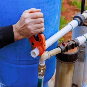 A close-up of a hand using a pipe cutter on a white PVC pipe, positioned next to a blue water tank and plumbing fixtures outdoors.