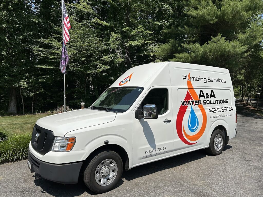 A white plumbing service van for A&A Water Solutions, featuring their logo with a flame and water droplet design, parked on a driveway surrounded by trees and American flags.
