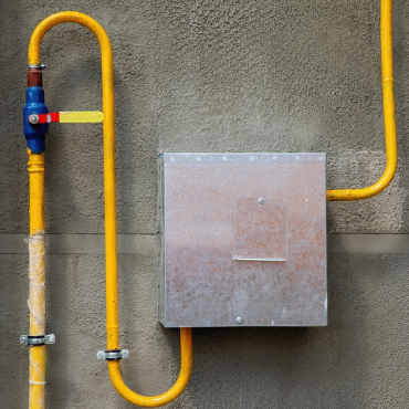 A yellow gas pipe with a valve and a protective metal box mounted on a gray wall, likely part of a gas line system installation.