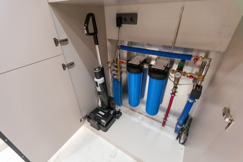 An image of a water filtration system installed inside a cabinet, featuring two blue filter cartridges and associated piping, with a vacuum cleaner placed nearby.