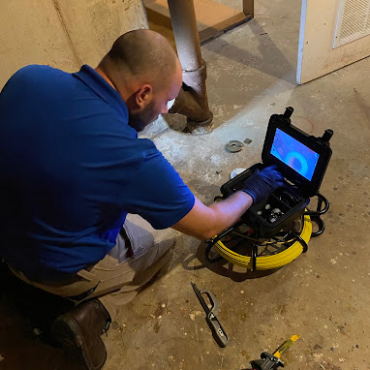 A plumber, wearing a blue shirt and gloves, kneels on the floor while operating a sewer inspection camera, viewing the footage on a monitor in a basement or utility area.